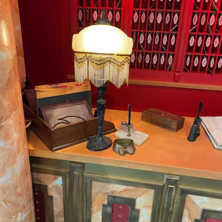 Illuminated vintage desk lamp with ornate base on an orange-toned desk, complemented by antique writing accessories and key cabinet in background.