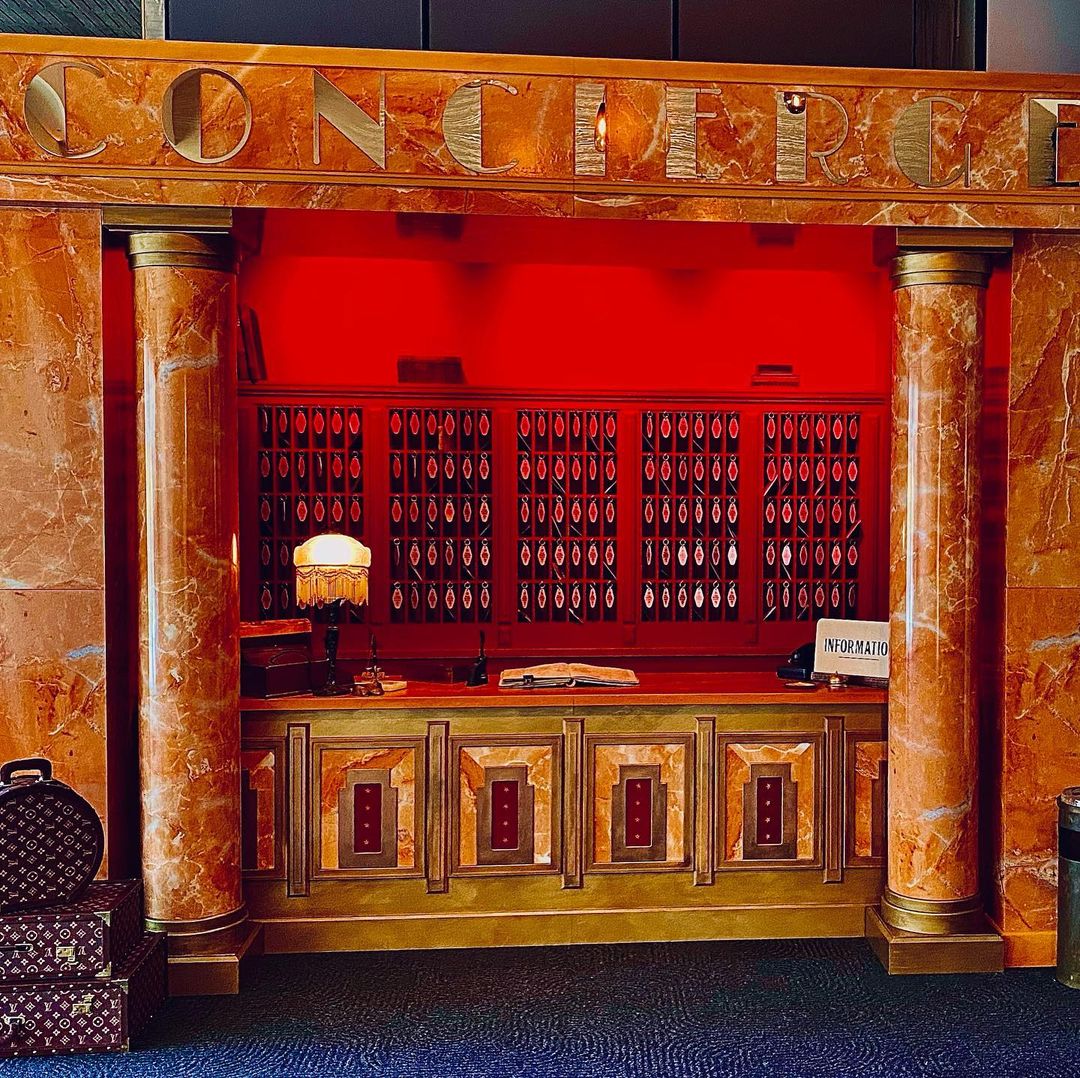 Grand hotel lobby with a marble reception desk, vintage-style lamp, and luggage. Red wall backdrop and architectural details.