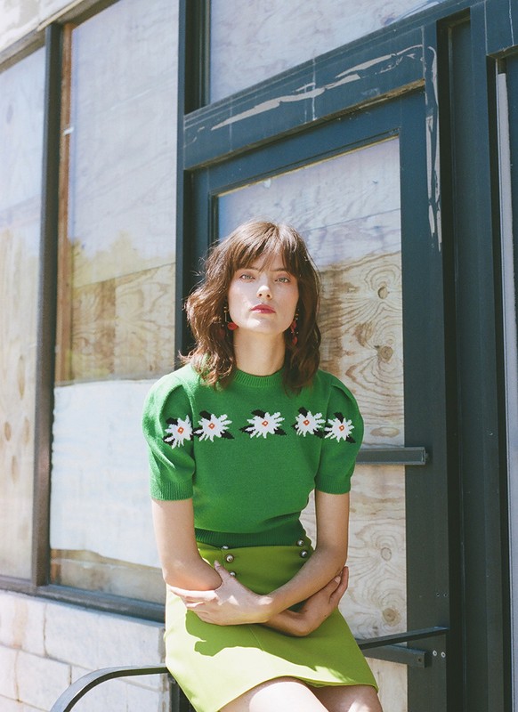 Adult woman in a green sweater with daisies and a lime green skirt, sitting against a weathered blue door with boarded up windows.