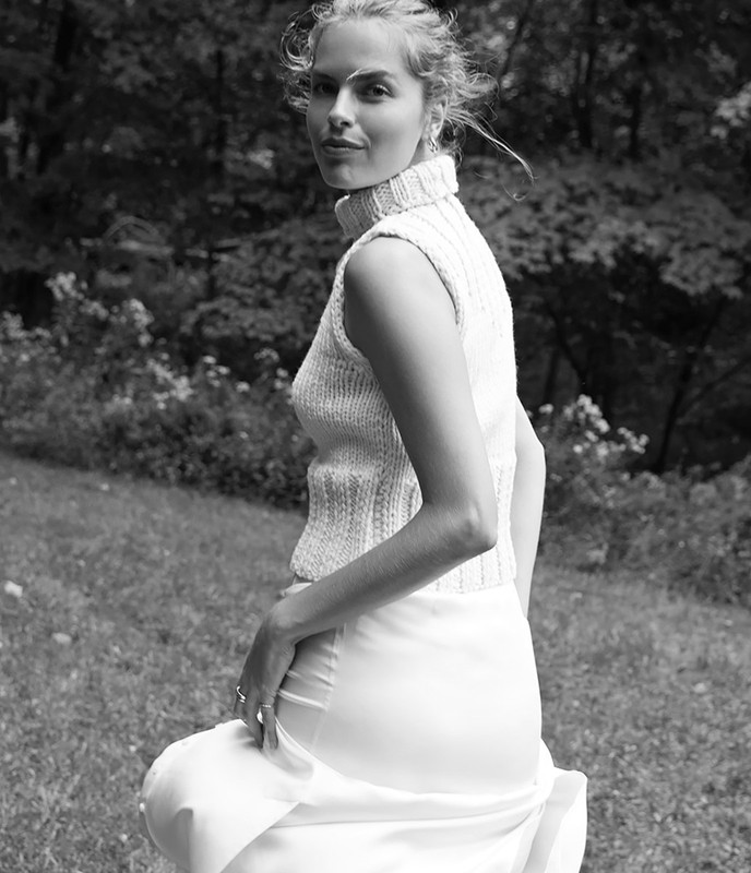 Black and white portrait of a woman in a knitted vest and long skirt standing in a grassy field with trees in the background.