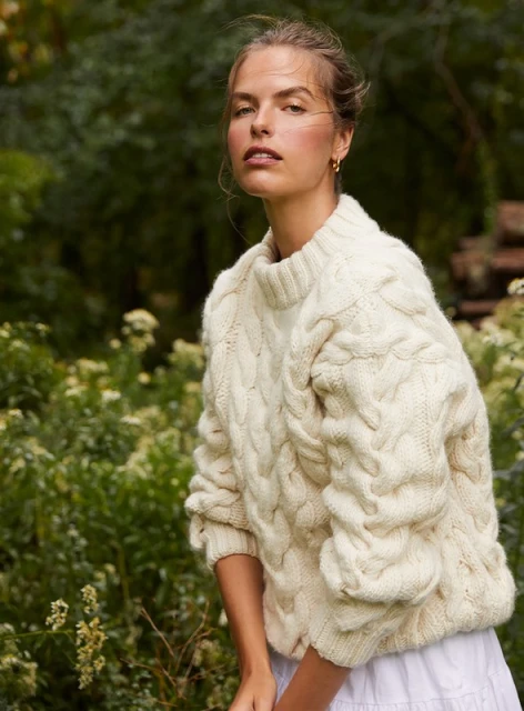 Portrait of a young adult woman wearing a cable knit sweater outdoors with a leafy green background in a natural setting.