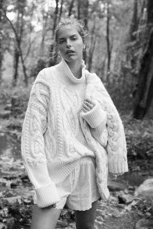 Monochrome portrait of a woman wearing a heavy cable-knit turtleneck sweater and shorts outdoors in a forest.