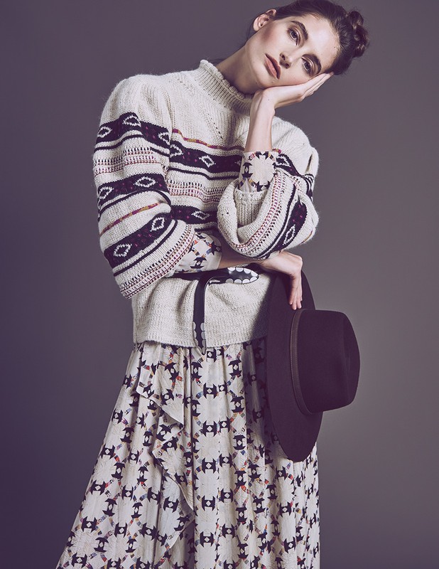 A fair-skinned young woman poses indoors wearing an patterned sweater and skirt, holding a fedora against a solid gray backdrop.