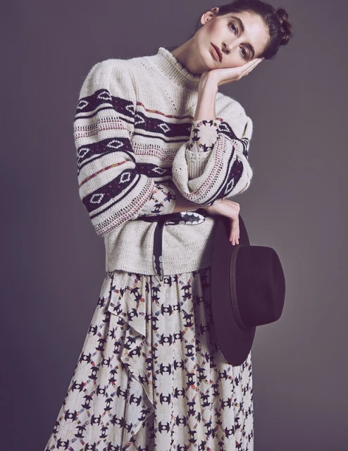 A fair-skinned young woman poses indoors wearing an patterned sweater and skirt, holding a fedora against a solid gray backdrop.