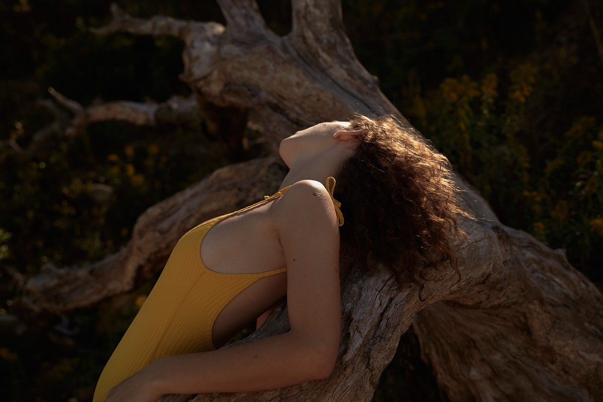 Young woman in yellow swimsuit rests against driftwood, eyes closed in the warm sunlight in a natural outdoor setting.