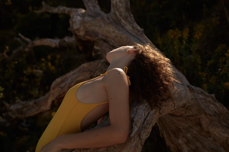 Young woman in yellow swimsuit rests against driftwood, eyes closed in the warm sunlight in a natural outdoor setting.