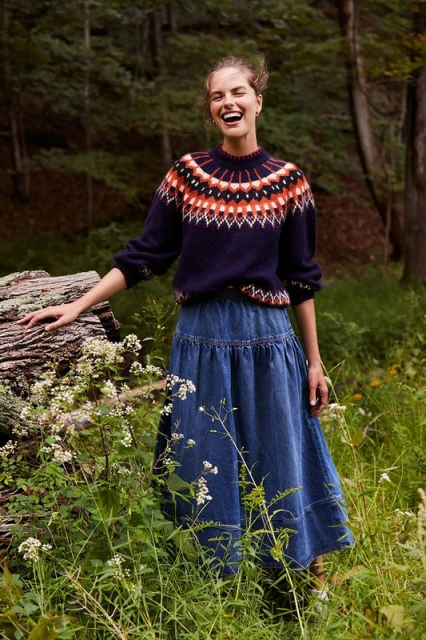A fair-skinned woman with a happy expression in a forest, wearing a patterned sweater and denim skirt, posing near a fallen tree.
