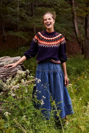 A fair-skinned woman with a happy expression in a forest, wearing a patterned sweater and denim skirt, posing near a fallen tree.