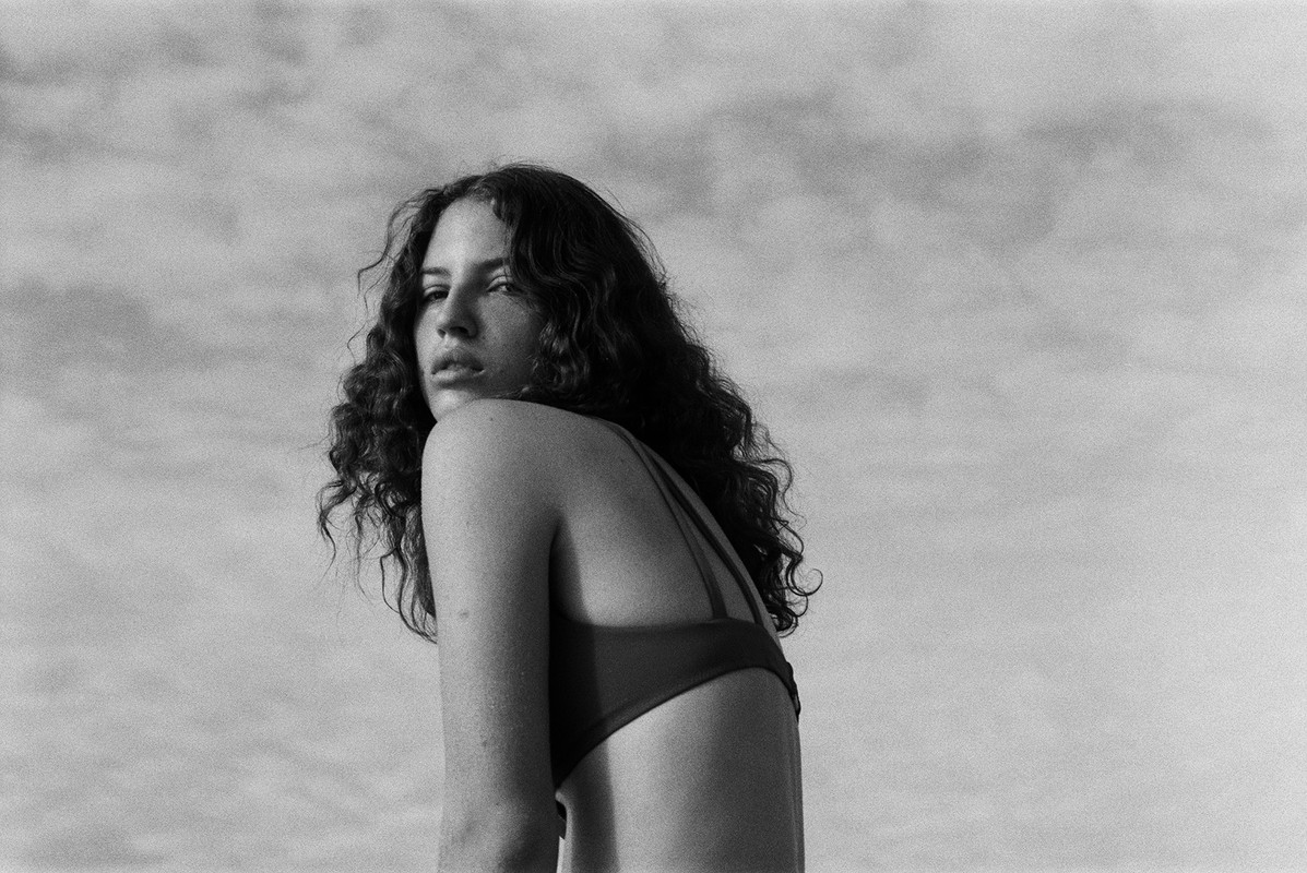 Black and white portrait of a woman with curly hair and a simple top, looking over her shoulder against a cloudy sky.