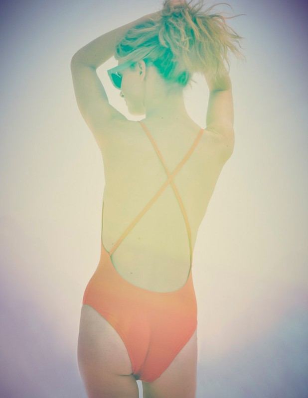 Woman in orange swimsuit with criss-cross straps stands with her back to the camera, arranging her blonde hair in an outdoor setting.