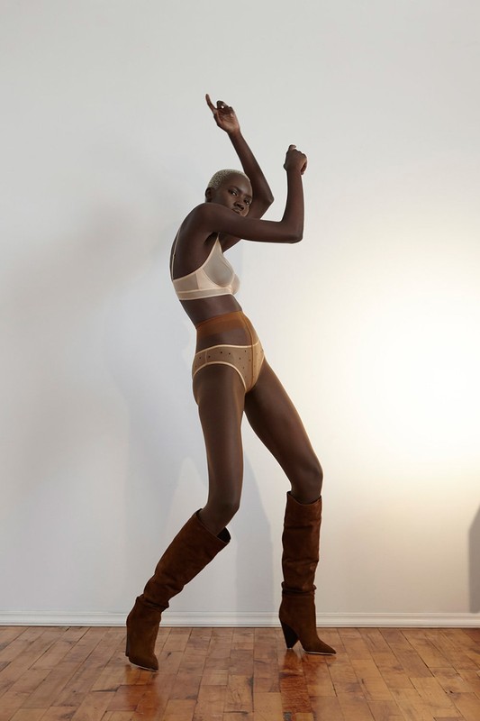 Full-length studio shot of a Black female model in underwear and boots, arms raised, against a neutral white background, with a wood floor.