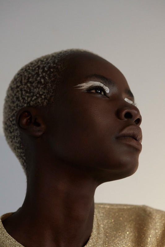 Close-up portrait of a stylish African woman with a white painted eyeliner design, light blonde hair, and a shimmering gold top.