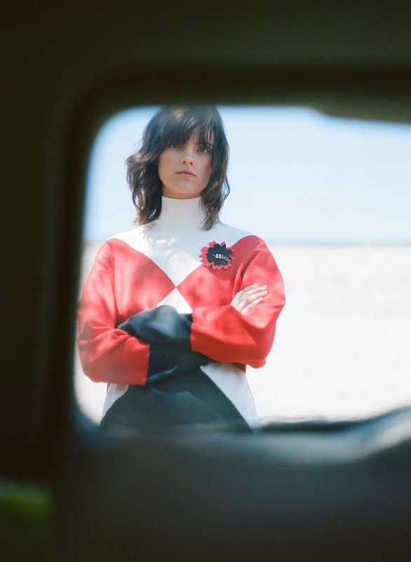 Fashion shot of a young woman with arms crossed in a red and white sweater, posing in an outdoor setting with a blurred window foreground.