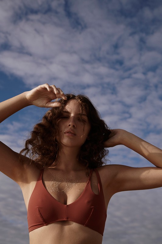 Low-angle portrait of a woman with curly brown hair wearing a rust-colored bikini top against a blue sky with clouds.