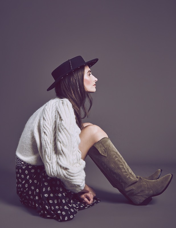A woman in a hat and sweater sits in a photo studio with cowboy boots. The scene is brightly lit against a gray backdrop.