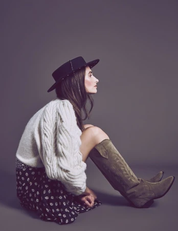 Profile view of a seated woman wearing a cream cable-knit sweater, dark hat, patterned skirt, and tall boots.