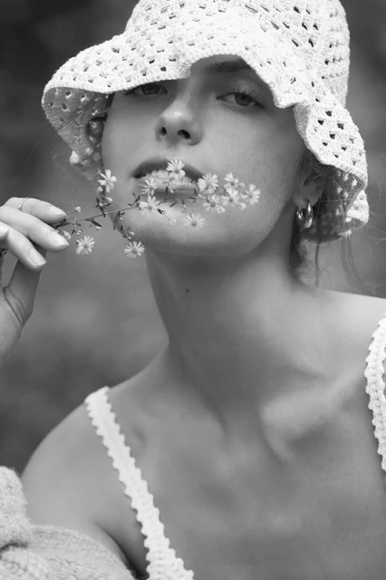 Black and white portrait of a young woman wearing a crocheted hat and holding a small sprig of flowers in her mouth outdoors.