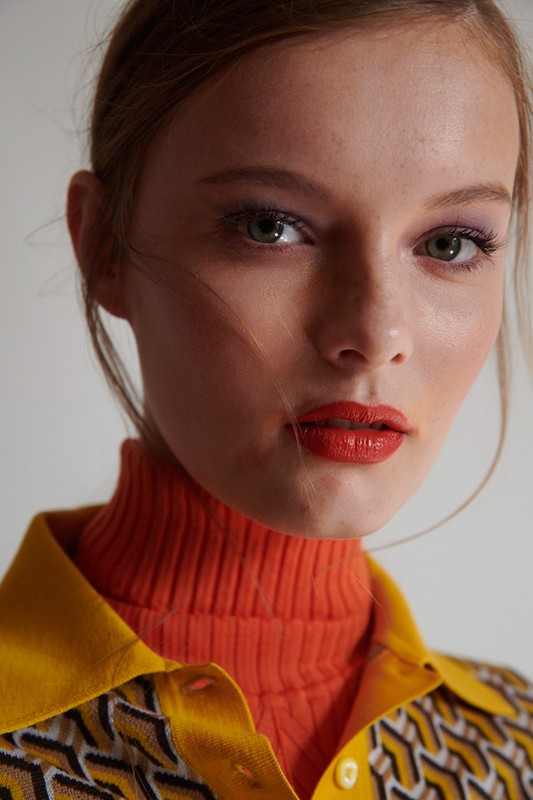 Close-up portrait of an adult female model with red lipstick and a patterned yellow shirt against a neutral background.