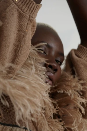Close-up portrait of a Black woman in a beige knit sweater and feathered collar with arms raised