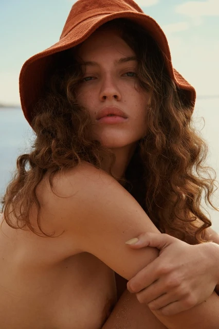 Close-up portrait of a woman wearing an orange hat, gazing at the camera with curly brown hair, against a blue and beige outdoor background.