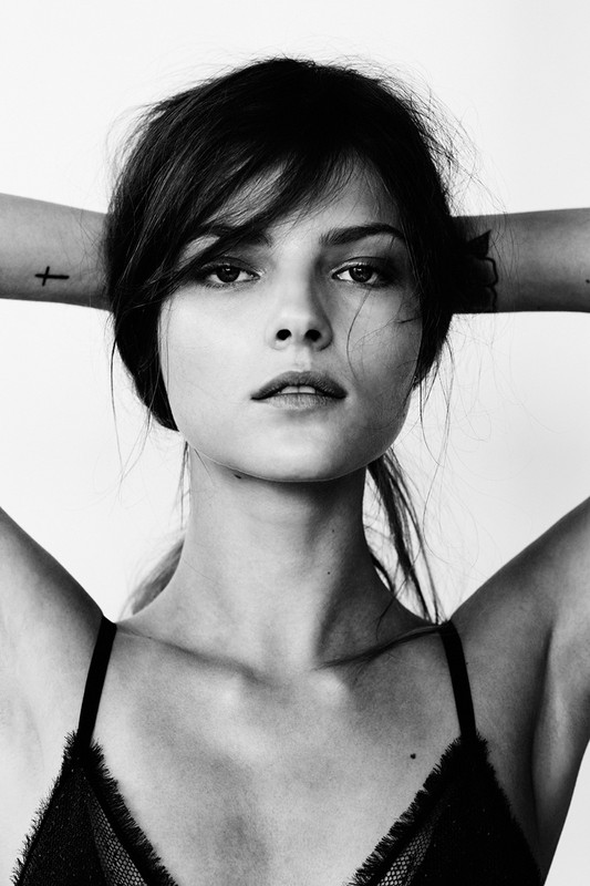 Portrait of a woman in a black lace top with her hands behind her head in front of a white wall in a monochrome studio shot.