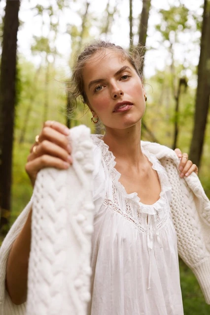 Portrait of a young woman in a white dress and cardigan in a forest. She is looking at the camera with a serious expression.