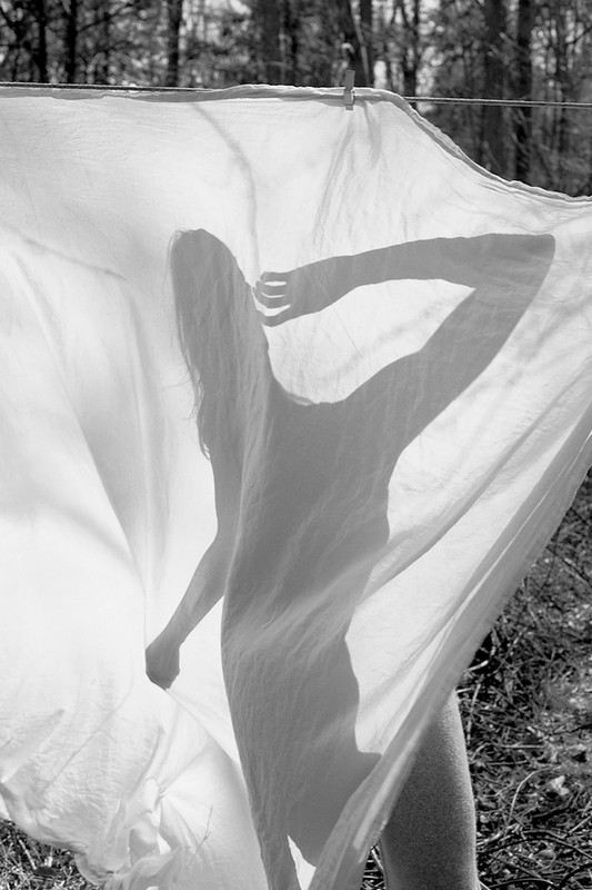 Silhouette of a woman visible through a white sheet hanging on a clothesline outdoors with trees in background.