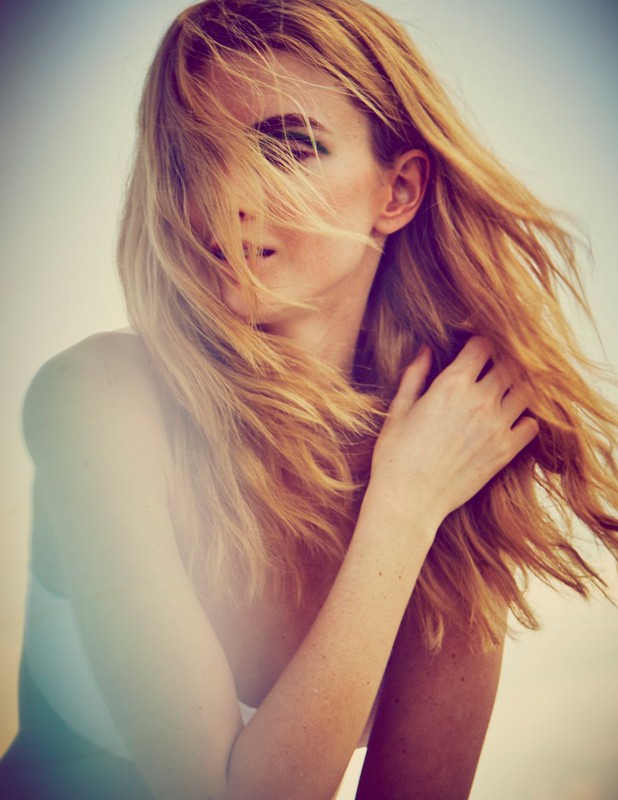 Portrait of a blonde woman with her hand in her hair, partially obscuring her face, in a bright outdoor setting with shallow depth of field.