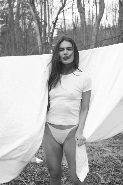 A woman poses outdoors in a white top and bottoms, holding fabric, with trees in the background. The image is black and white.