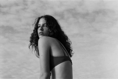 Monochrome portrait of a woman with curly hair looking back over her shoulder against an overcast sky