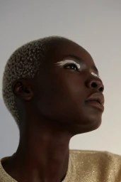 Close-up portrait of a Black woman with short blonde buzzcut and white graphic eyeliner wearing a metallic gold top
