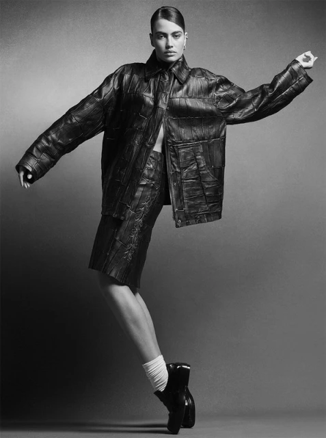Studio shot of a female model in an oversized leather jacket and shorts, posing in black heeled boots against a neutral background.