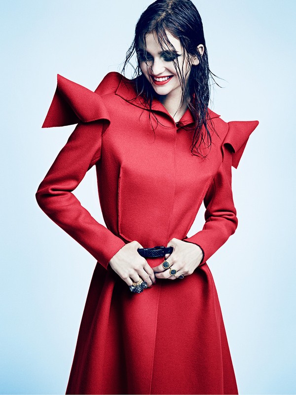 Model with wet dark hair wearing a structured red coat with statement shoulders and red lipstick against a light blue background