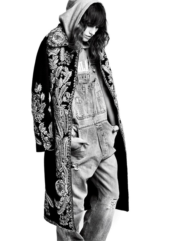 Black and white portrait of a woman wearing a hooded sweatshirt, denim overalls, and a long patterned coat, standing in a bright setting.