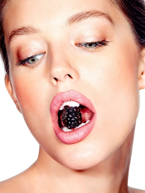 Close-up of a woman's face with light skin and pink lips, holding a blackberry in her mouth, against a white background.