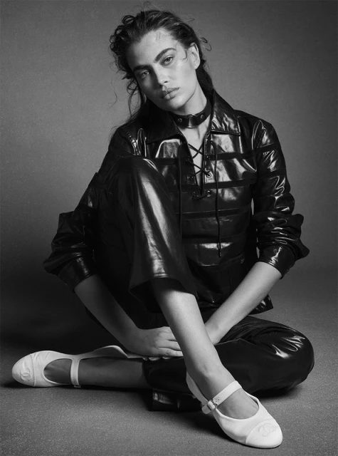 Black and white portrait of a woman in a shiny jacket, leather-look pants, and white shoes, sitting indoors against a gray backdrop.