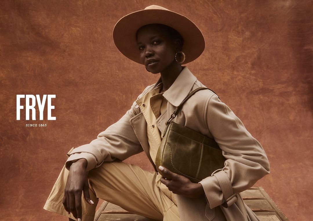 Dark-skinned woman wearing a tan hat and trench coat poses with a green purse in front of an earth-toned background, showcasing fashion.
