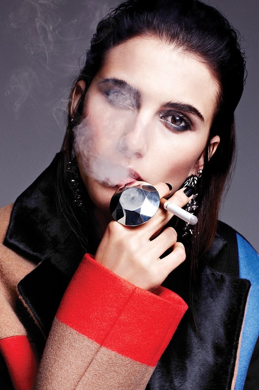 Stylish woman with dark hair, dark eyes, heavy makeup, and red lipstick smoking a cigarette wearing an angular ring and jacket.
