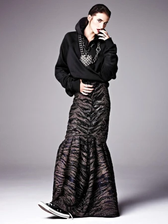 Model in a black hoodie and patterned maxi skirt wearing chunky chain jewelry against a grey studio backdrop