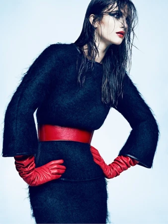 Model with wet hair wearing a black textured dress, red belt, and red leather gloves against a pale blue background.