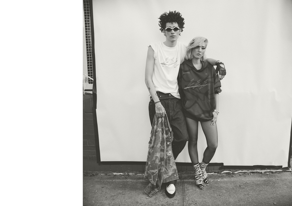 A monochromatic fashion-style photo showcasing a male and female standing close together in stylish outfits in front of a grey wall.