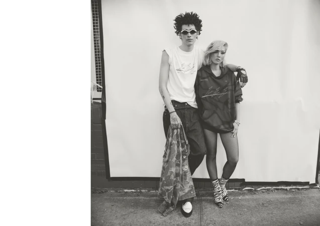 A monochromatic fashion-style photo showcasing a male and female standing close together in stylish outfits in front of a grey wall.