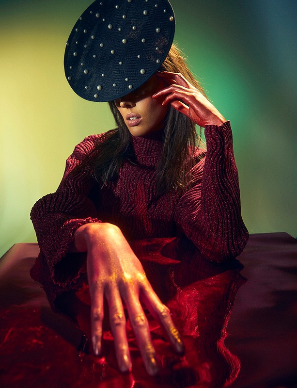 Portrait of a woman with a studded hat and gold painted hands in a studio with blended green and yellow lighting.