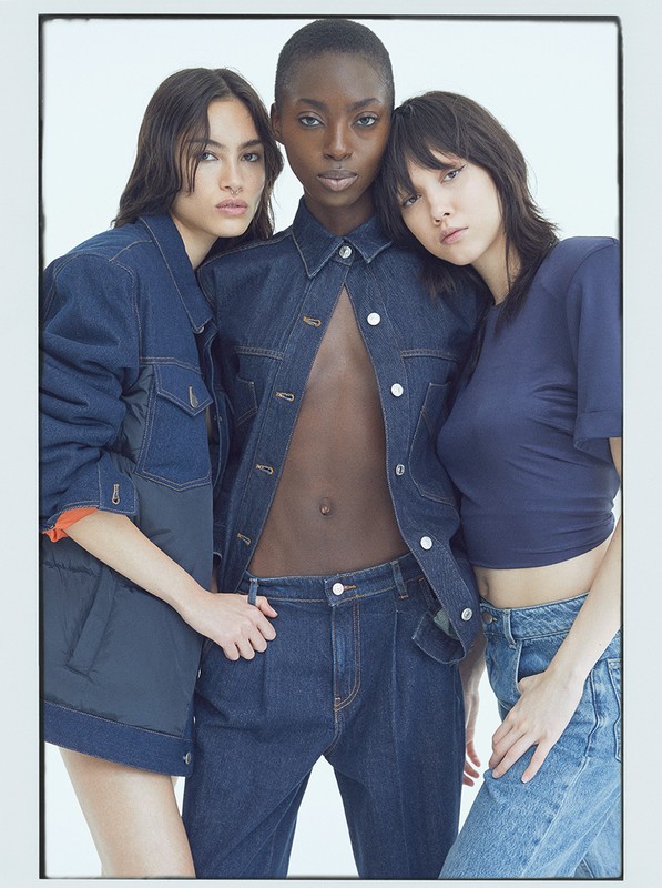 Three women pose indoors wearing denim jackets and jeans against a white backdrop in a fashion studio shot with soft, diffused lighting.