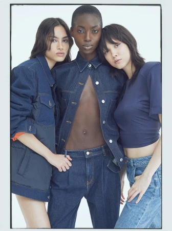 Three women pose indoors wearing denim jackets and jeans against a white backdrop in a fashion studio shot with soft, diffused lighting.