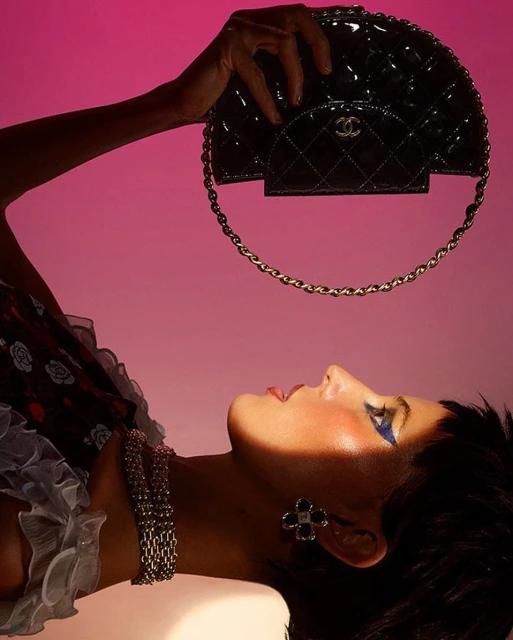 Adult female laying down in a studio setting with pink background, holding a glossy Chanel half-moon handbag above her head.