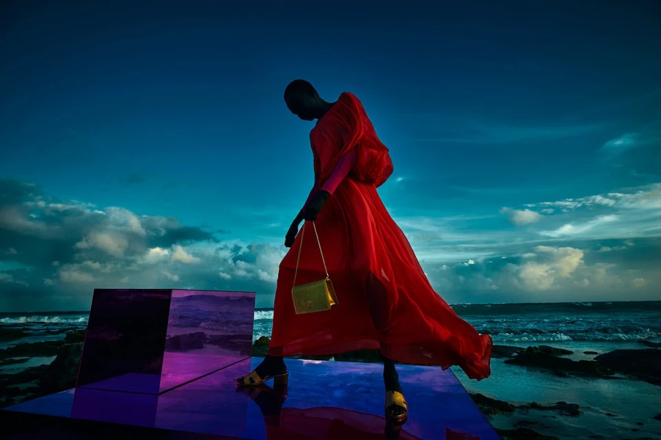 A dark-skinned fashion model in a red dress and handbag walks on a reflective platform before an ocean and sky at dusk.