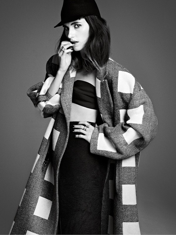 Black and white portrait of a woman in a hat and coat, in a studio setting, wearing black skirt and top with hand near her mouth.