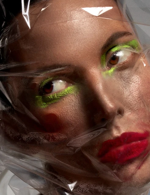 Close-up portrait of a woman with bright green eyeshadow and smeared red lipstick covered in clear plastic wrap for beauty shot.