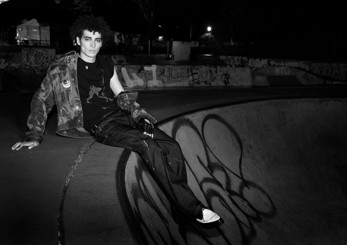 Black-and-white portrait of a young adult male with dark curly hair wearing a jacket, t-shirt, and jeans, sitting on a concrete structure outdoors.